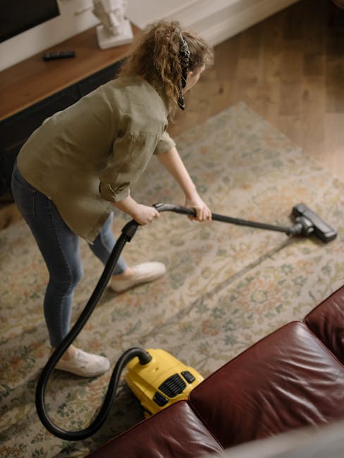 A professional cleaner from Mayfair Carpet Cleaners, dressed in a white protective suit, blue gloves, and a face mask, is performing deep cleaning on a beige carpet in a modern, well-lit living room. 