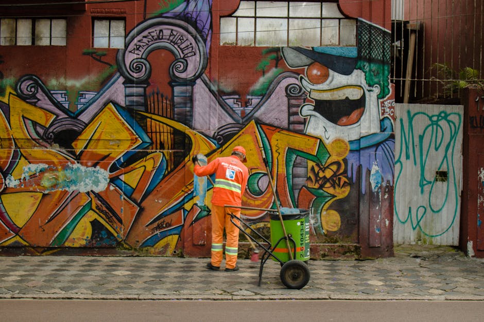 Two individuals wearing orange overalls are engaged in graffiti removal on a grey stone wall along a sidewalk near Piccadilly and Mayfair. One person, with glasses and dark hair tied back, holds a spr