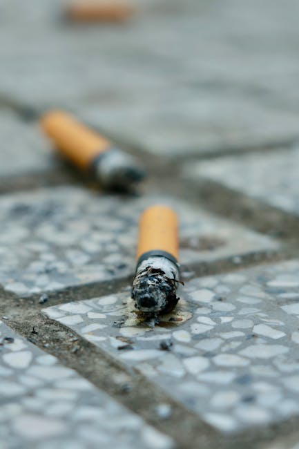 Close-up view of two discarded cigarette butts on a tiled outdoor surface with small pebble insets, with one butt in focus showing ash and burnt cigarette filter, while the other is slightly blurred i