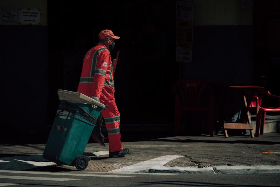 A street scene with a person dressed in a dark raincoat and hood, standing next to a green trash bin on wheels, which is tucked into a plastic liner. The individual appears to be collecting waste or p